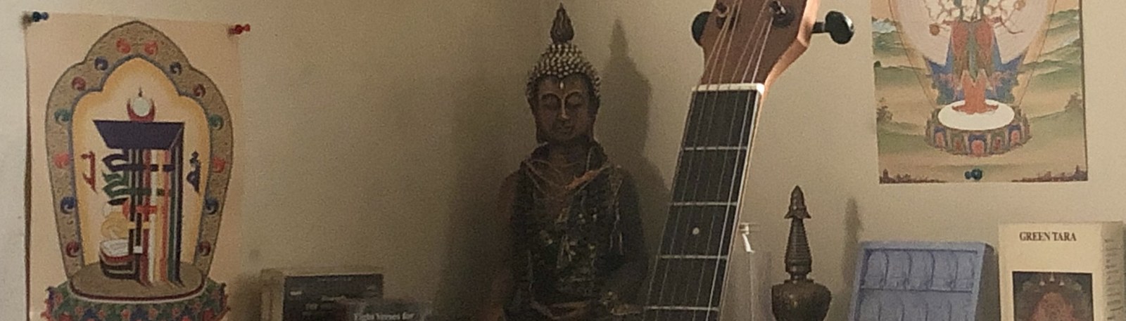 A home altar in low light: a seated Buddha between hanging thangkas, a brass stupa, a lit candle and the neck of a guitar.