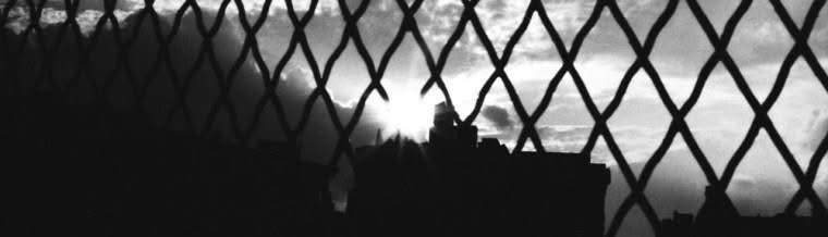 A city skyline in silhouette behind a chain-link fence, sun breaking low through the diamonds of the wire.
