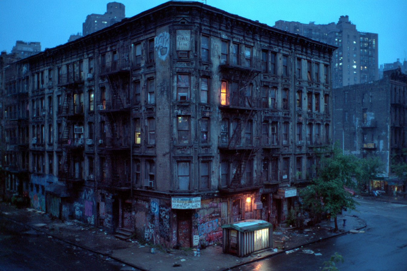 Squat Politics featured: a graffiti-covered corner tenement with fire escapes at dusk, a lit newsstand on the corner.